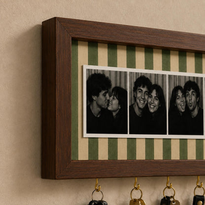 A rectangular dark wood wall-mounted key holder featuring a central horizontal strip of green and beige vertical stripes. The frame displays a series of three black and white photographs of a couple, with four brass hooks attached below.