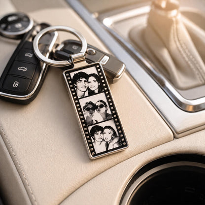 A rectangular metal keychain featuring a vertical film strip design with three black and white photographs of two people. The keychain is attached to a silver metal ring and is positioned next to a black car key fob.