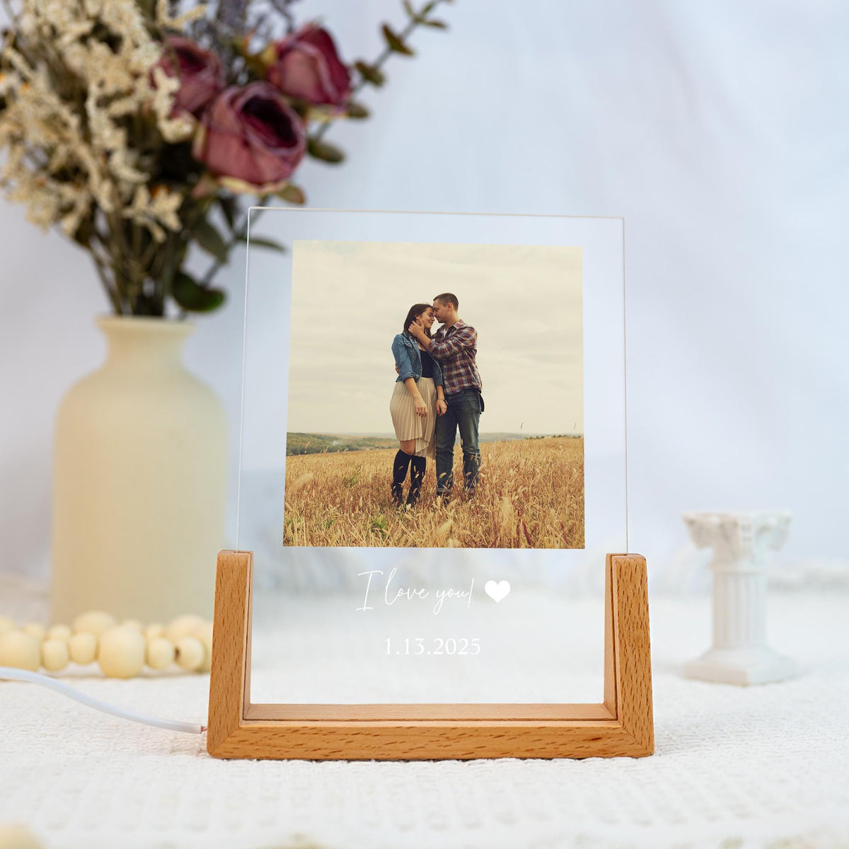 A clear acrylic photo frame with a wooden base, displaying a couple in a field. The text "I love you!" and the date 1.13.2023 are printed on the frame. A vase of flowers is in the background.