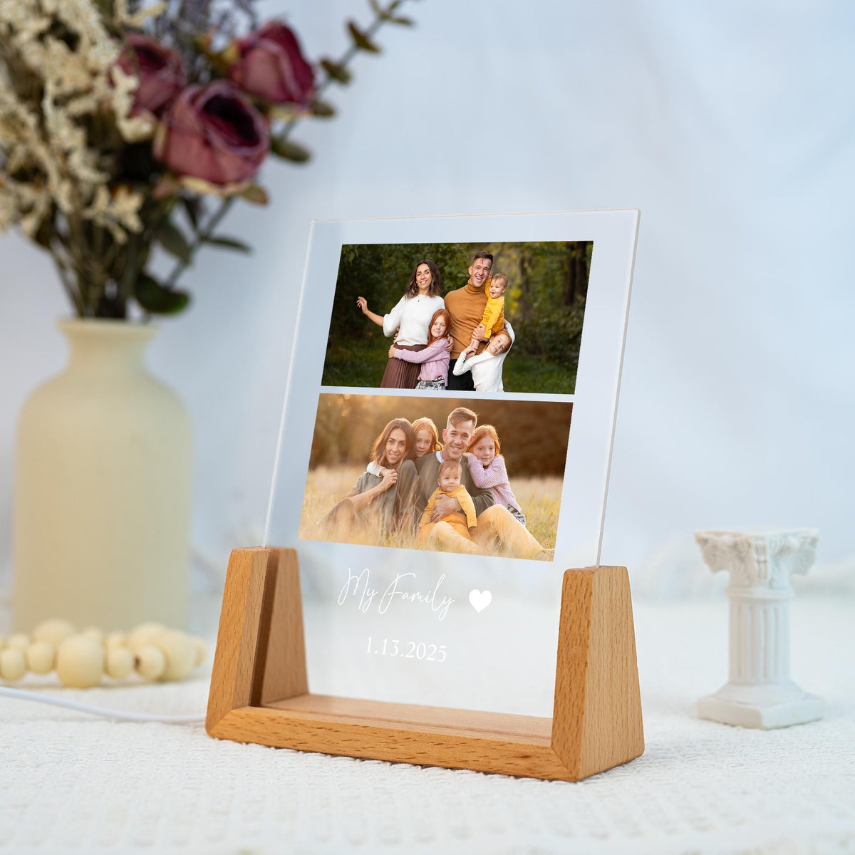 A clear acrylic photo display with two family photos, set in a wooden base. The text "My Family" and the date 1.13.2025 are printed on the acrylic. A vase of flowers and a small white decorative column are in the background.