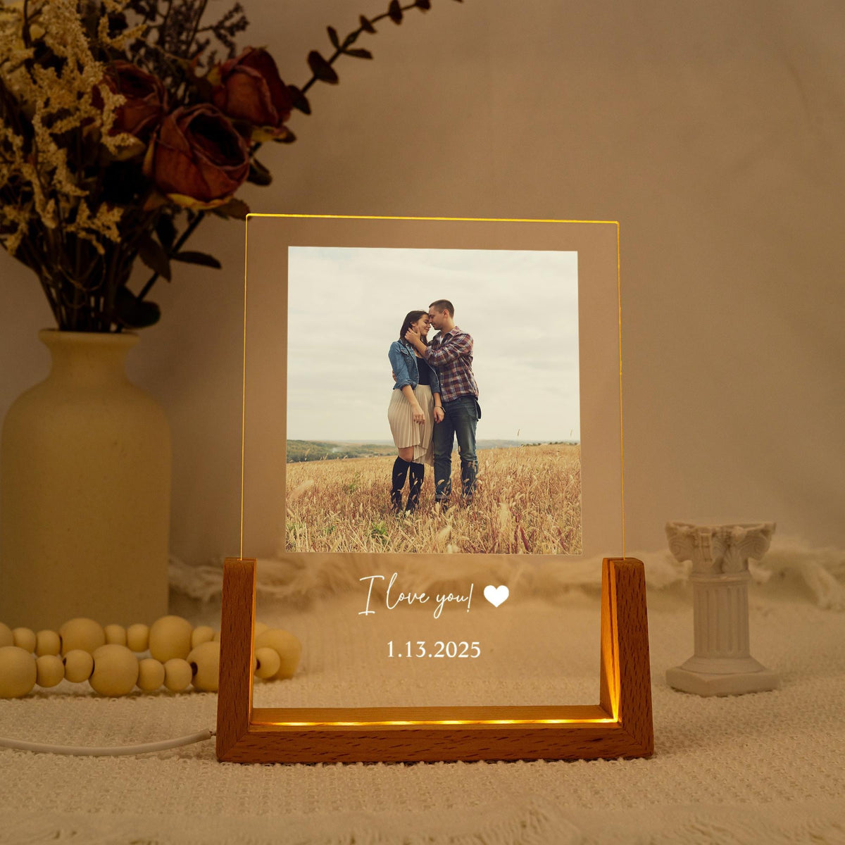 A lighted photo frame with a wooden base. The frame displays a couple in a field and the text "I love you!" with a heart symbol and the date 1.13.2025. The frame is illuminated with a warm glow.