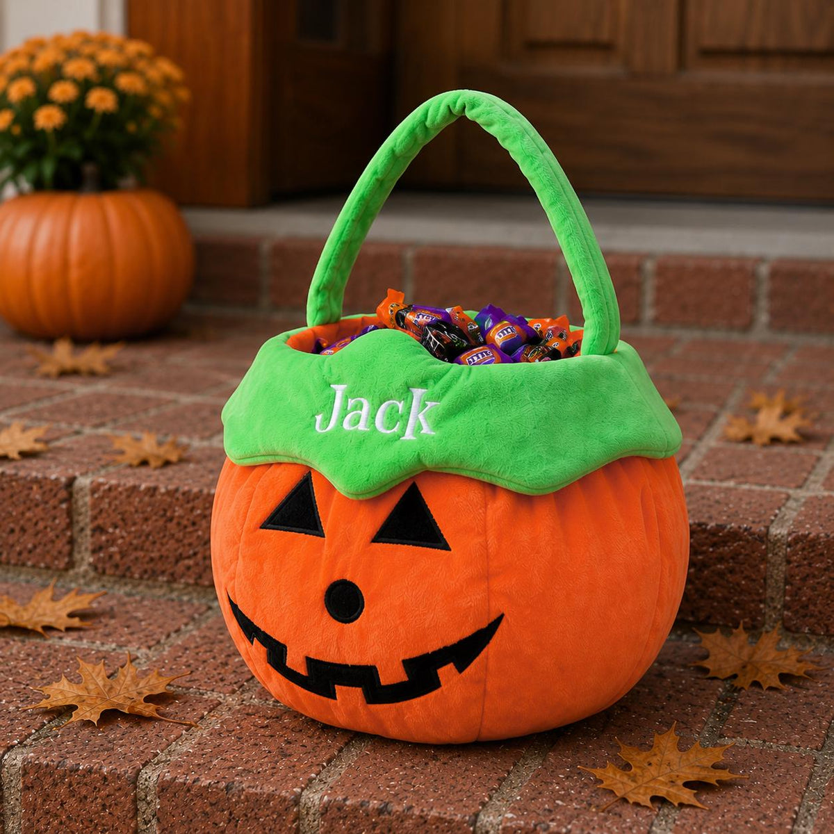 A plush orange pumpkin-shaped Halloween candy bucket with a green handle and trim. The front features a black felt jack-o'-lantern face and the name Jack embroidered in white thread on the green rim. The bucket is filled with candy.
