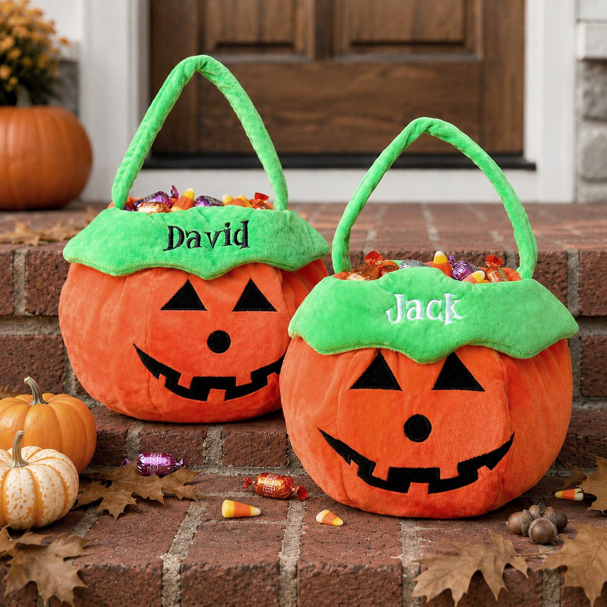 Two plush orange pumpkin-shaped candy buckets with green handles and scalloped rims. The left bucket features the name David embroidered in black, and the right bucket features the name Jack embroidered in white. Both have black jack-o'-lantern faces.