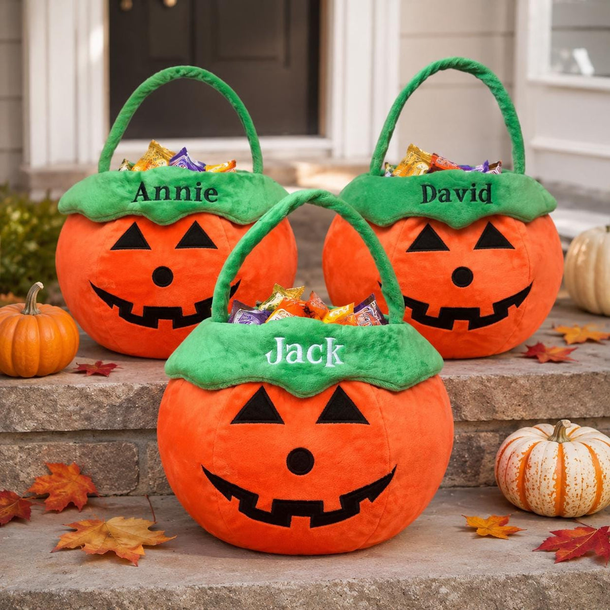 Three plush orange pumpkin-shaped Halloween candy buckets with green handles and rims. Each bucket features a black jack-o'-lantern face and is personalized with embroidered names: Annie, David, and Jack in white or black thread.