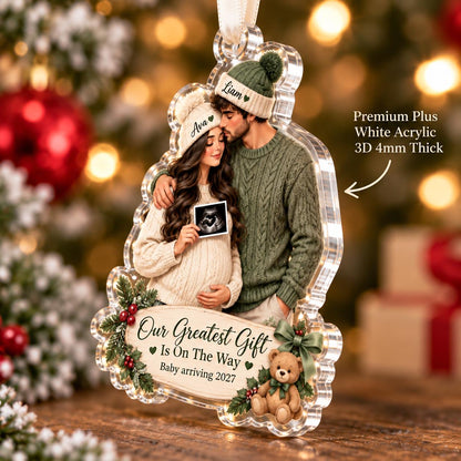 May include: A clear acrylic ornament featuring a couple wearing knit hats and sweaters, holding a sonogram. The ornament includes the text "Our Greatest Gift Is On The Way" and "Baby arriving 2027," decorated with holly, berries, and a teddy bear. It is 4mm thick.