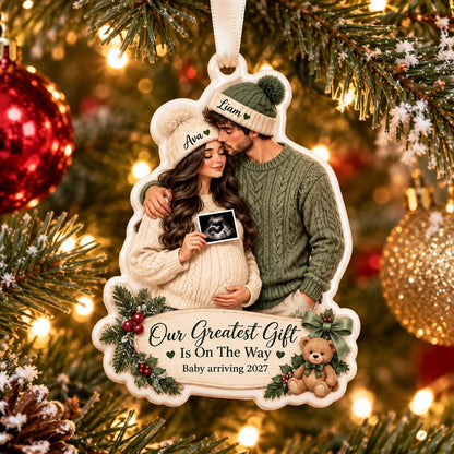May include: A white, shaped Christmas ornament featuring an illustration of a couple wearing knit hats and sweaters, holding an ultrasound image. The text reads "Our Greatest Gift Is On The Way" and "Baby arriving 2027" with a small teddy bear.