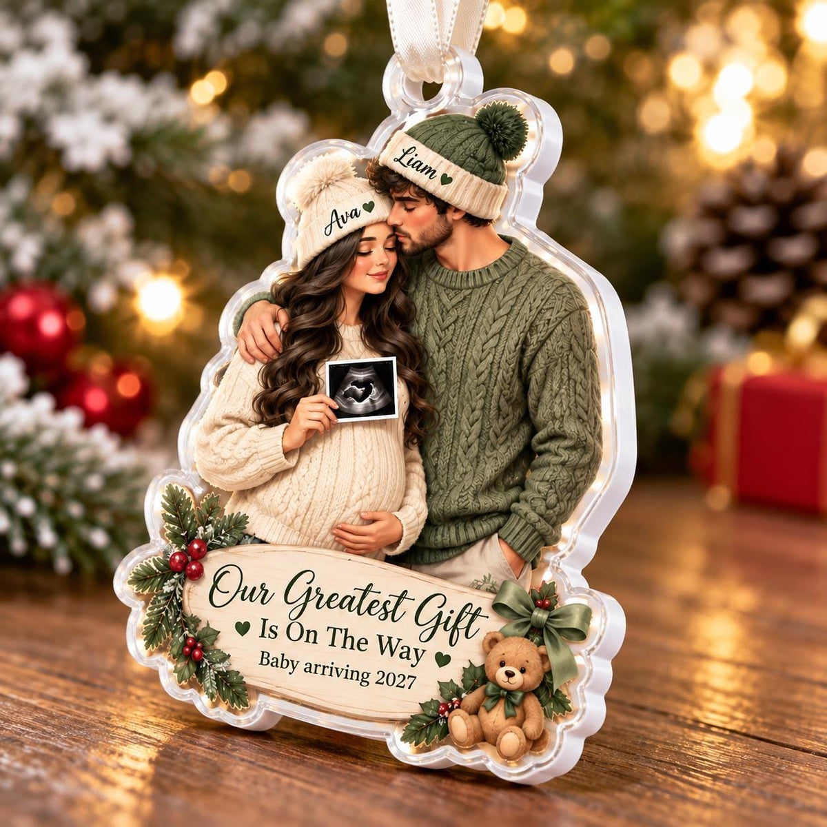 May include: A clear acrylic Christmas ornament featuring an illustration of a couple wearing matching green beanies labeled Ava and Liam. The woman holds an ultrasound image. Text reads Our Greatest Gift Is On The Way Baby arriving 2027.