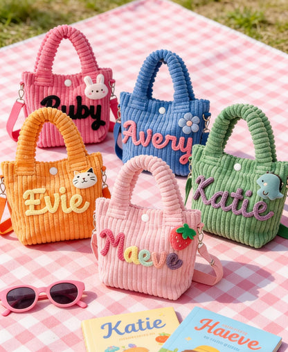 Five colorful corduroy tote bags with names embroidered on them. The bags are pink, blue, orange, light pink, and green. Each bag has a handle and a shoulder strap. The bags are decorated with a bunny, a flower, a cat, a strawberry, and a sea creature.