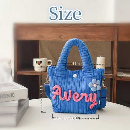 Blue corduroy tote bag with the name "Avery" in pink script. The bag has a flower accent and a shoulder strap. It measures 11 inches wide and 8.3 inches high.