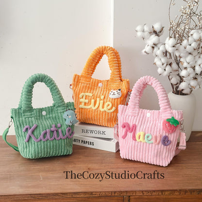 Three corduroy tote bags in green, orange, and pink, each with a name embroidered on the front. The green bag has a blue dinosaur, the orange bag has a cat, and the pink bag has a strawberry. The bags have handles and shoulder straps.
