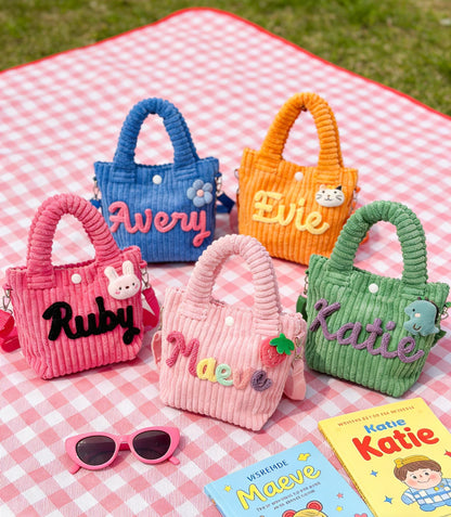 Five colorful corduroy tote bags with names embroidered on them. The bags are in shades of blue, orange, pink, light pink, and green. Each bag has a matching handle and a small decorative charm. Pink sunglasses and two books are in the foreground.