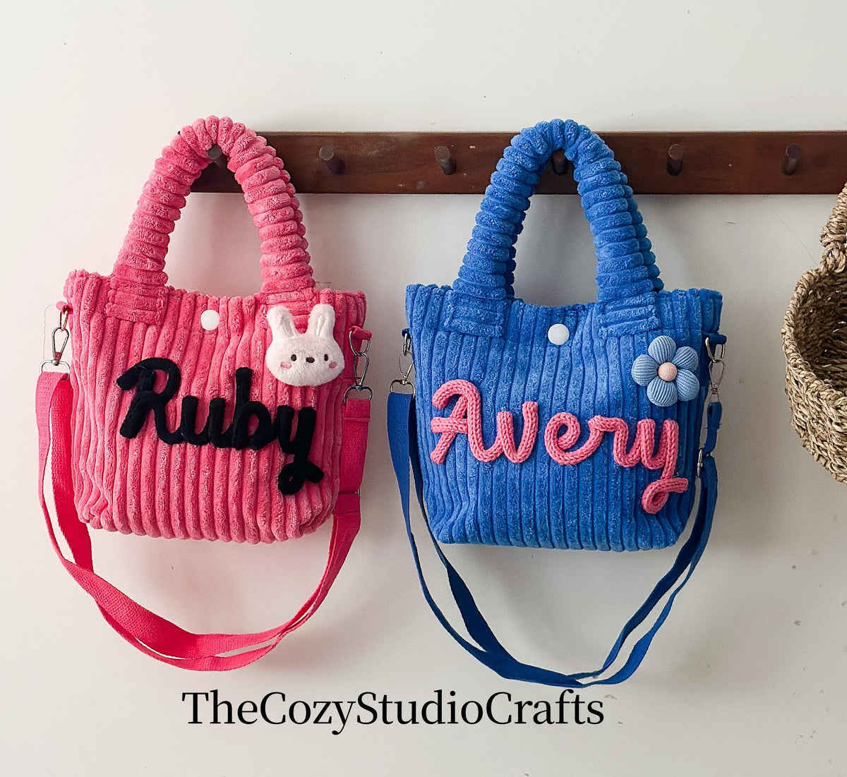 Two corduroy tote bags, one pink and one blue, each with a name embroidered on the front. The pink bag has the name "Ruby" in black and a white bunny. The blue bag has the name "Avery" in pink and a blue flower.