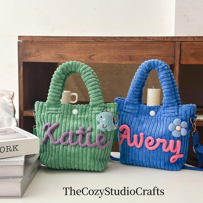 Two corduroy tote bags, one green and one blue, with matching handles. The green bag has the name "Katie" in purple, and the blue bag has the name "Avery" in pink. Each bag has a small charm.