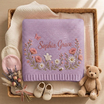 A lavender knitted baby blanket featuring the embroidered name Sophia Grace surrounded by pink and white floral motifs, a butterfly, and a bee. The set includes two small knitted hats, a pair of baby shoes, and a small teddy bear.