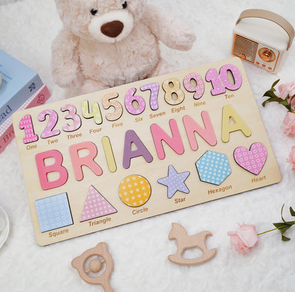 A rectangular wooden puzzle board featuring the name BRIANNA in large, colorful letters, numbers one through ten, and various shapes including a square, triangle, circle, star, hexagon, and heart, each labeled with its name.