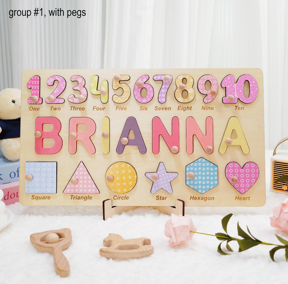 A wooden puzzle board featuring the text group #1, with pegs. The board displays numbers one through ten, the name BRIANNA, and shapes including a square, triangle, circle, star, hexagon, and heart, each with a small wooden peg.