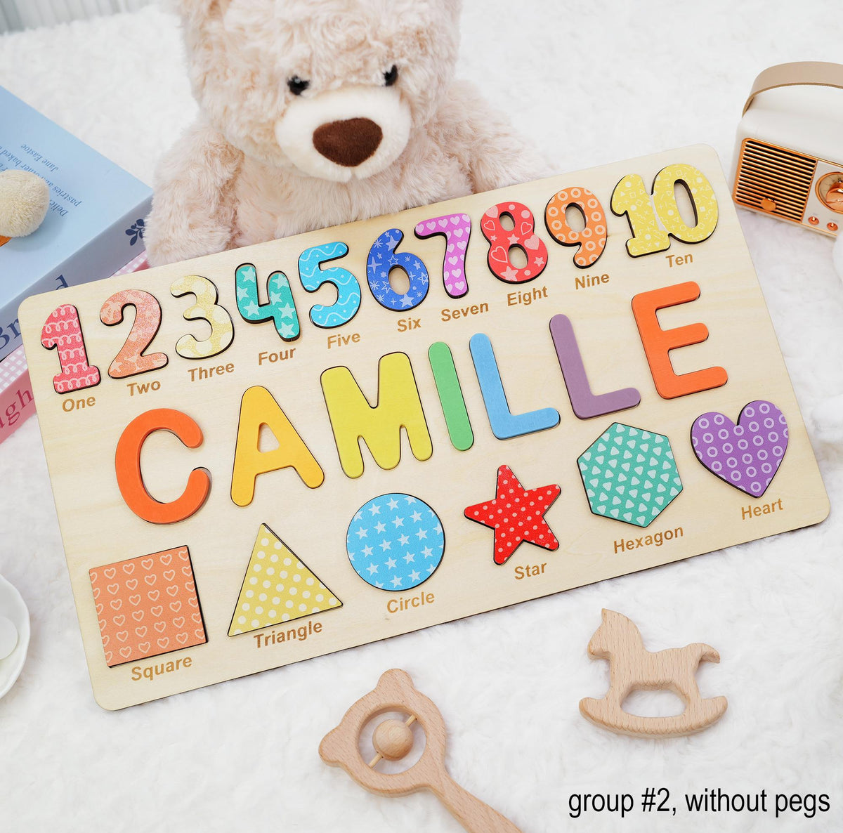 A wooden puzzle board featuring the name CAMILLE in large colorful letters, numbers one through ten with corresponding words, and various shapes including a square, triangle, circle, star, hexagon, and heart with labels.
