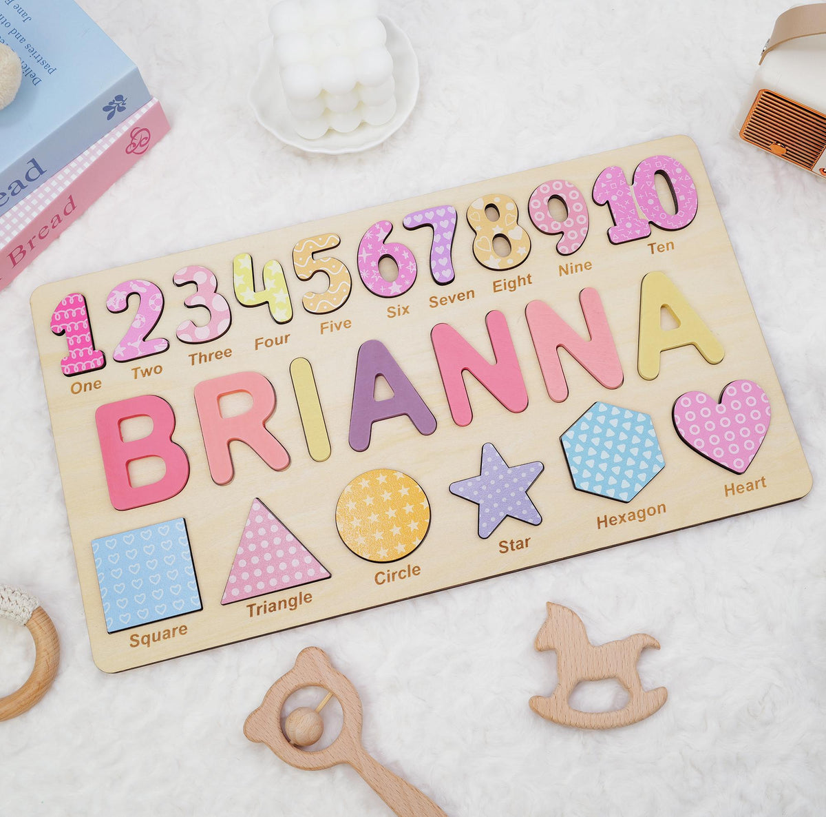 A rectangular wooden puzzle board featuring the name BRIANNA in large, colorful letters, numbers one through ten with corresponding labels, and various shapes including a square, triangle, circle, star, hexagon, and heart.