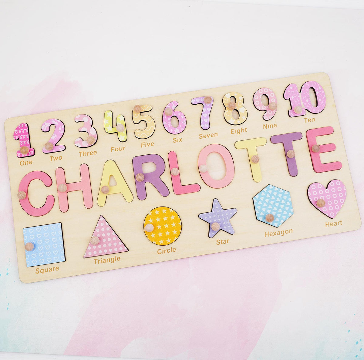 A rectangular wooden puzzle board featuring the name CHARLOTTE in large, colorful letters, numbers one through ten, and various shapes including a square, triangle, circle, star, hexagon, and heart, each with a small wooden knob.