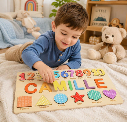 A wooden educational puzzle board featuring the name CAMILLE in large, colorful letters, a row of numbers from one to ten with patterned designs, and a bottom row of geometric shapes including a square, triangle, circle, star, hexagon, and heart.