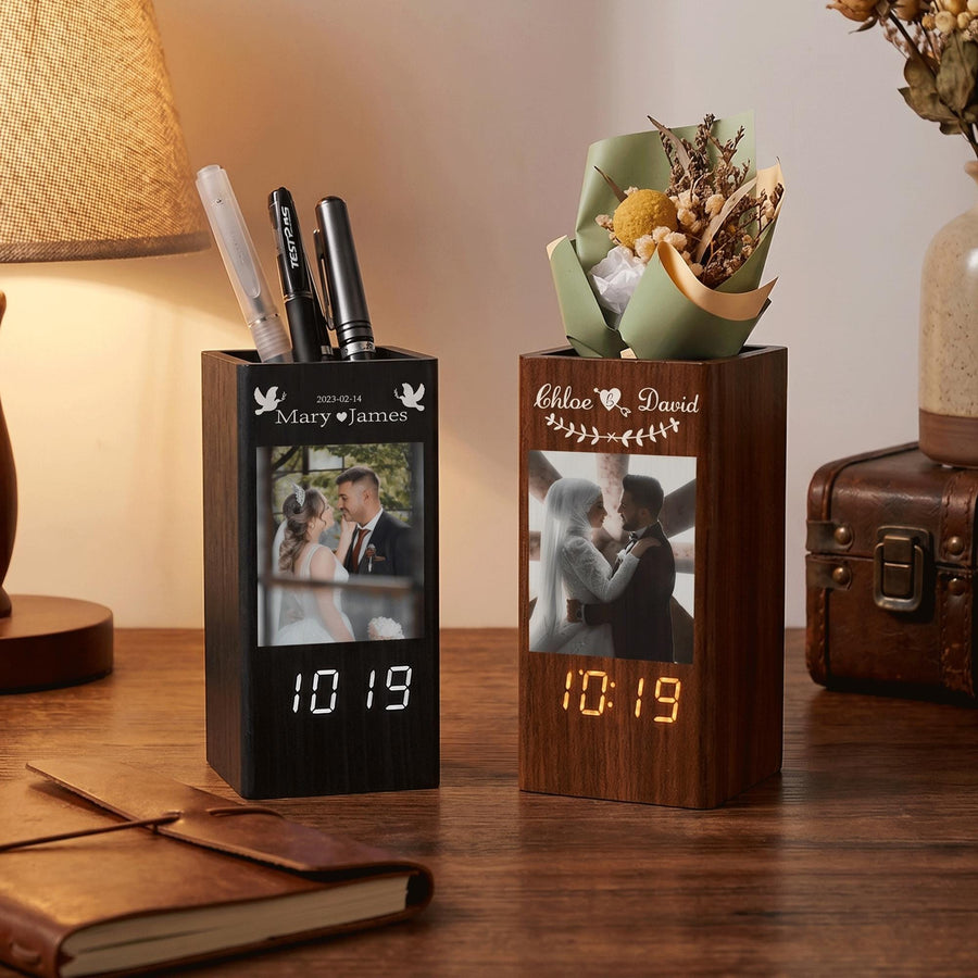 Two rectangular wooden desk organizers with integrated digital clocks. The left organizer is dark wood with a photo of a couple, the date 2023-02-14, and two pens. The right organizer is medium wood with a photo of a couple and dried flowers.