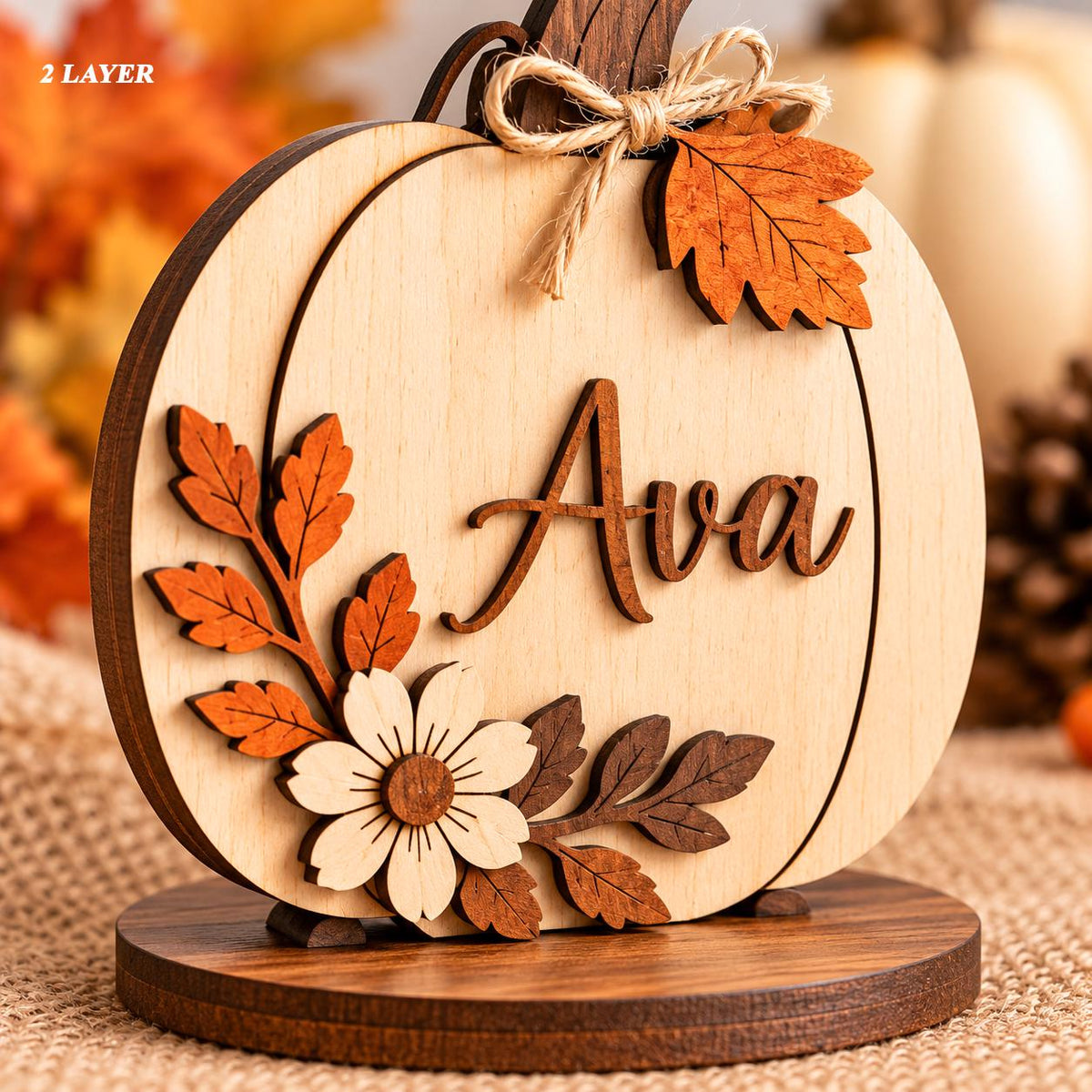 May include: A two-layer wooden pumpkin decoration featuring the name Ava in dark brown script. The pumpkin is adorned with a light-colored wooden flower, dark brown and orange wooden leaves, and a small jute twine bow on the stem.