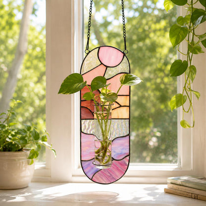 A stained glass window panel in shades of pink, orange, and purple, with a black metal frame and chain for hanging. A small glass vase holds a green plant. The panel is hanging in front of a window.