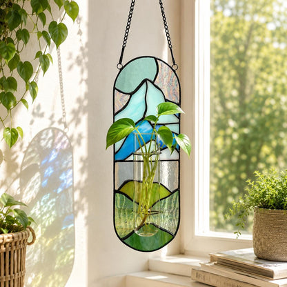 A stained glass hanging planter with a black metal frame. The glass features a mountain scene in shades of blue, green, and white. A clear glass tube holds a green plant with large leaves. The planter hangs from a black chain.