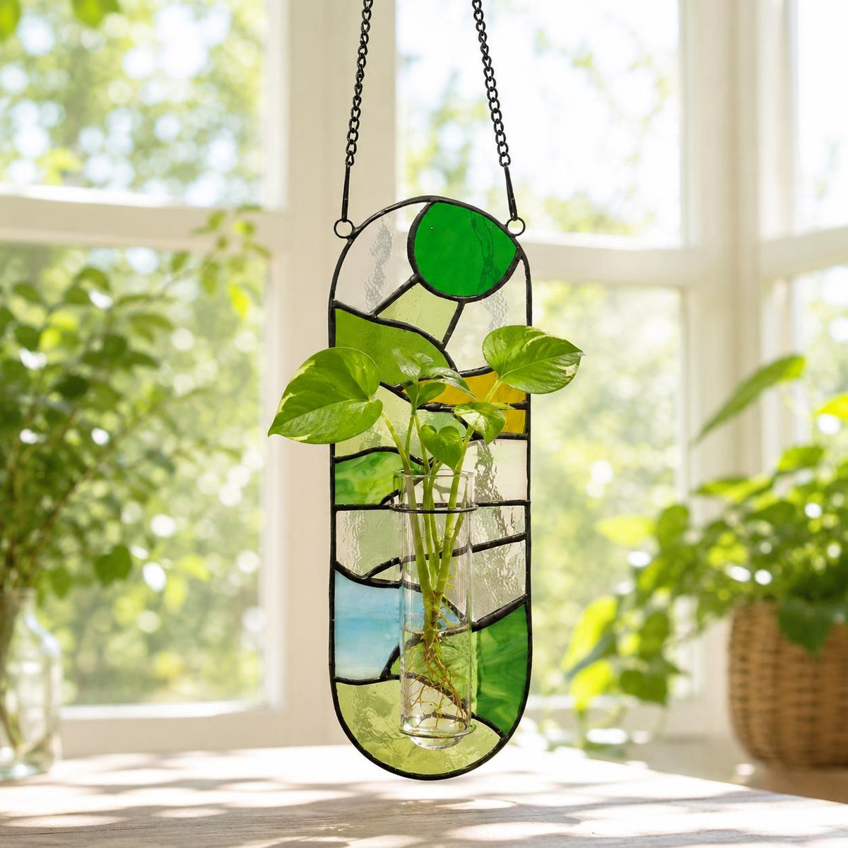 A stained glass plant holder with a clear glass tube for plants. The holder is a vertical oval shape with green, yellow, and blue glass pieces. It hangs from a black chain. The plant has green leaves.