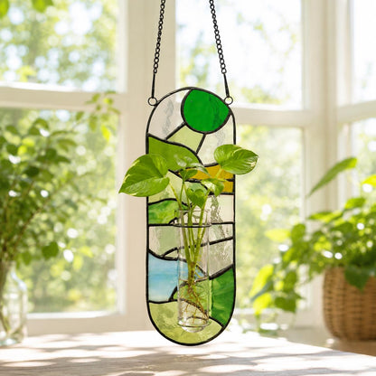 A stained glass plant holder with a clear glass tube for plants. The holder is a vertical oval shape with green, yellow, and blue glass pieces. It hangs from a black chain. The plant has green leaves.