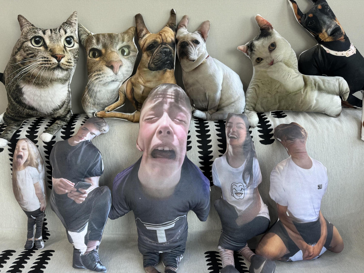 A collection of personalized pillows featuring various faces, including cats, dogs, and people. The pillows are arranged on a couch with a black and white patterned fabric.