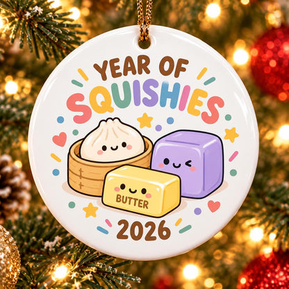 May include: A round white ceramic ornament featuring the text YEAR OF SQUISHIES and 2026. The design includes a cartoon dumpling in a bamboo steamer, a yellow butter block labeled BUTTER, and a purple cube, all with smiling faces and colorful accents.